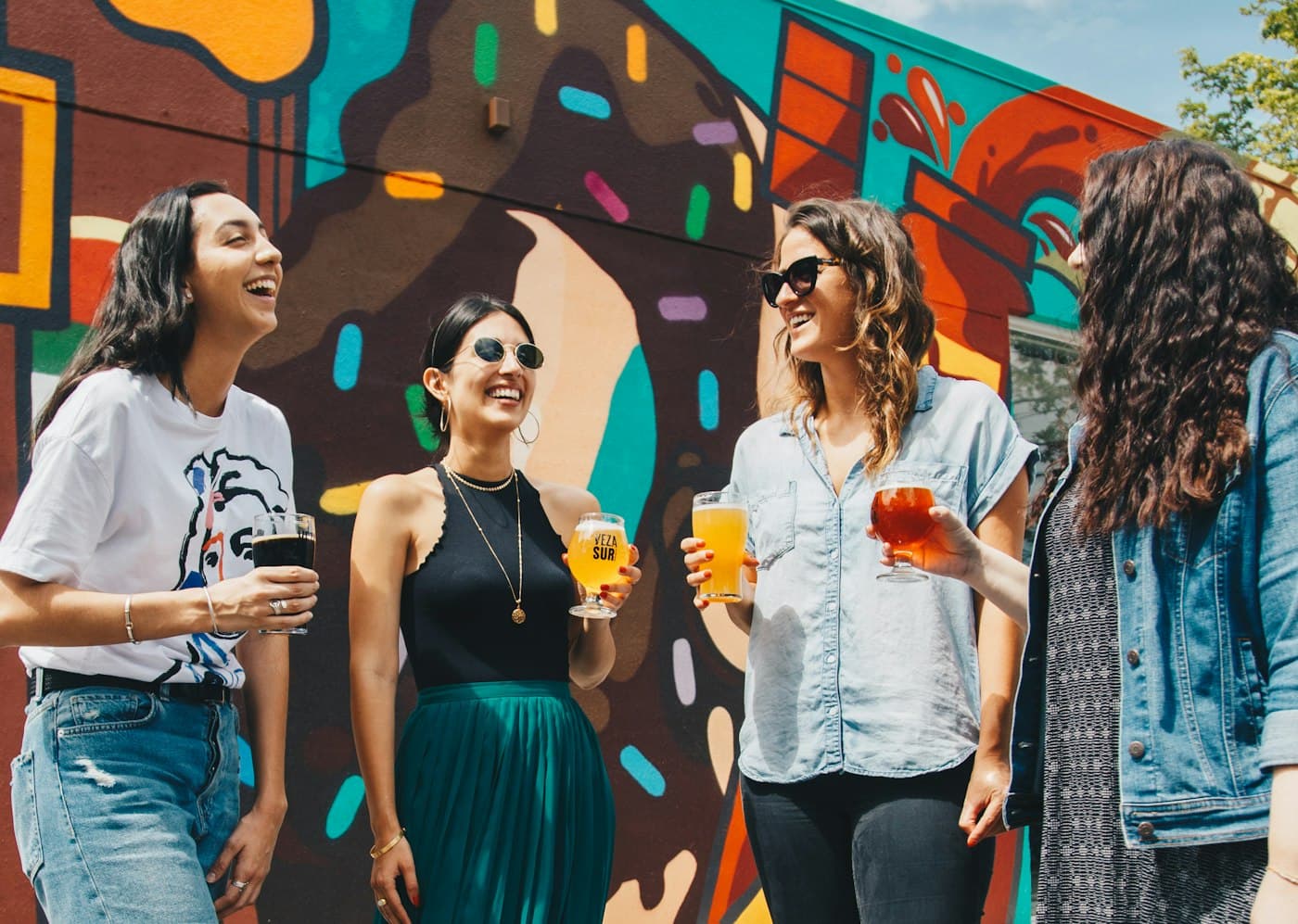 Four friends laughing together with drinks in front of a city mural